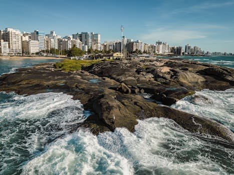 pexels-photo-15138937-15138937 A scenic view of Guarapari's beachfront with crashing waves and city skyline in the background.
